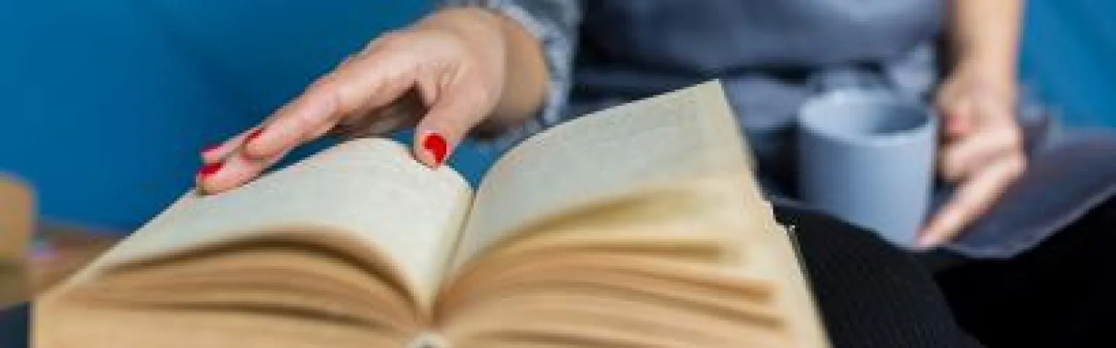 Book being read by a women with red nails holding a coffee cup.